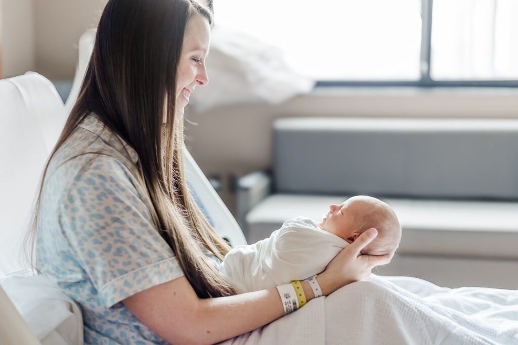 Mom smiling while holding newborn baby boy swaddled in white blanket during fresh-48 session at UAB in Birmingham, AL 