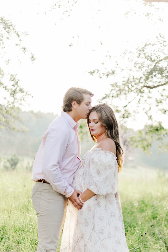 mother looking down with her partner kissing her head in a serene boho field, with golden sunlight filtering through the trees.