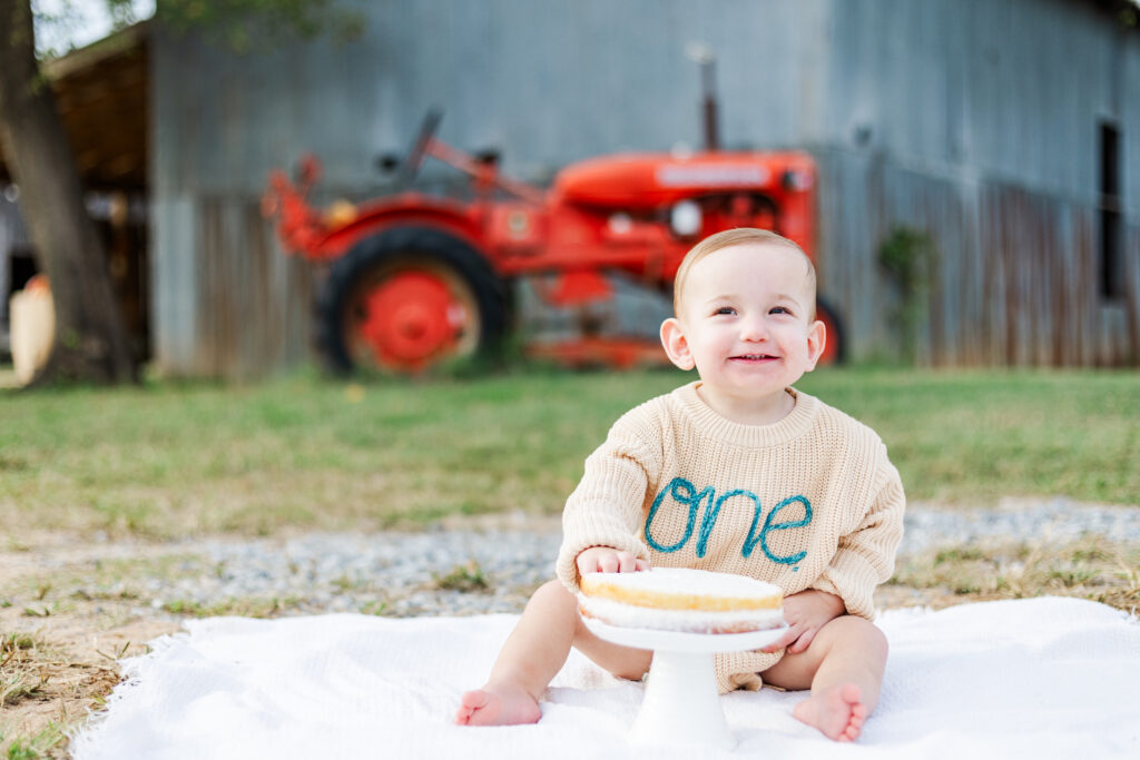 A joyful toddler sitting in front of a white smash cake at Old Baker Farm, surrounded by rustic farm decor during smash cake session