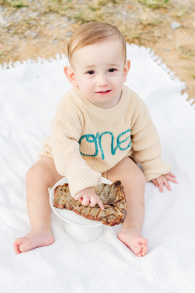 Little boy sitting on white blanket with t-bone steak during 12 month milestone session with 5U Photography