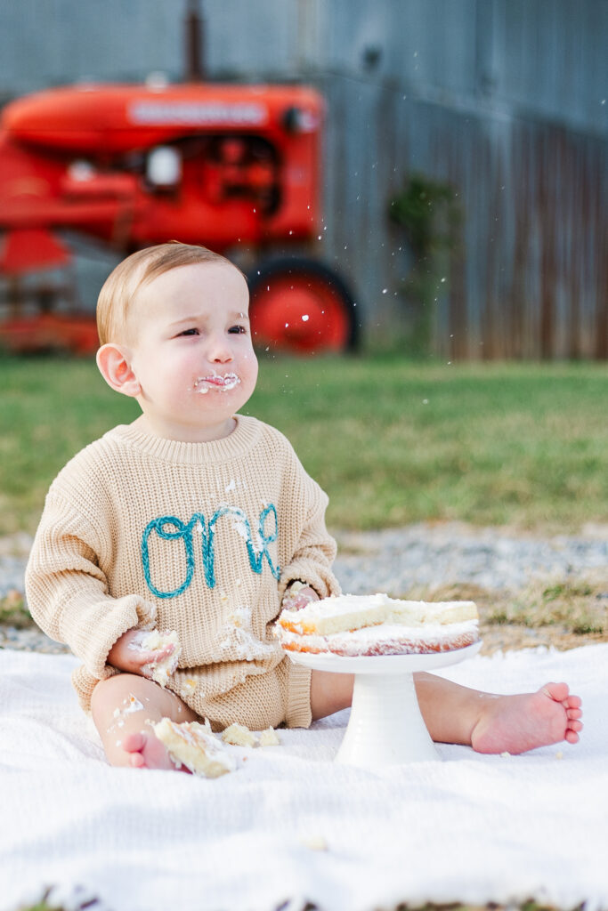 Little boy sitting on white blanket in front of tractor at Old Baker Farm during smash cake session with 5U Photography 