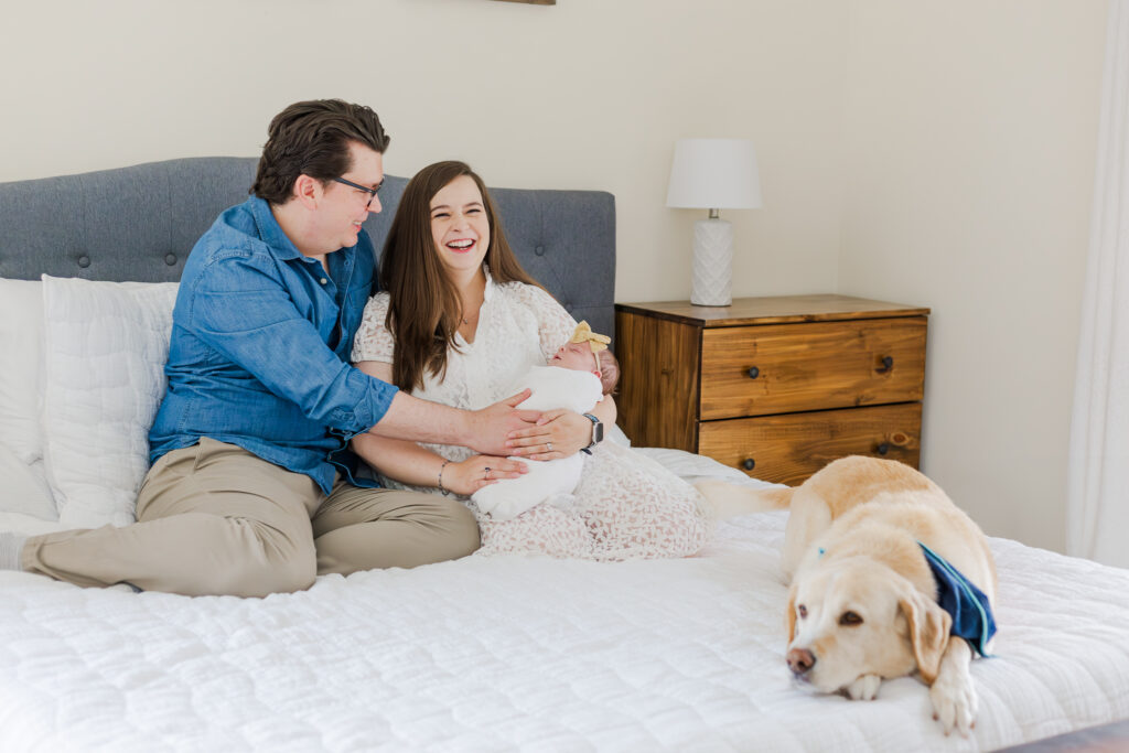 Mom and dad sitting on bed holding newborn baby girl laughing at camera during in-home newborn session in birmingham alabama