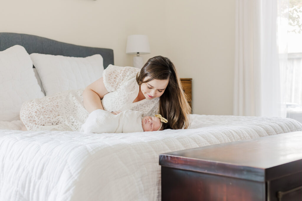 Mom lying on bed looking down at newborn baby girl swaddled in white blanket during in-home lifestyle newborn session in birmingham alabama with 5u photography
