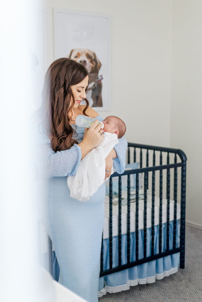 Mom holding baby boy feeding him a bottle in front of his crib during newborn session in hoover alabama