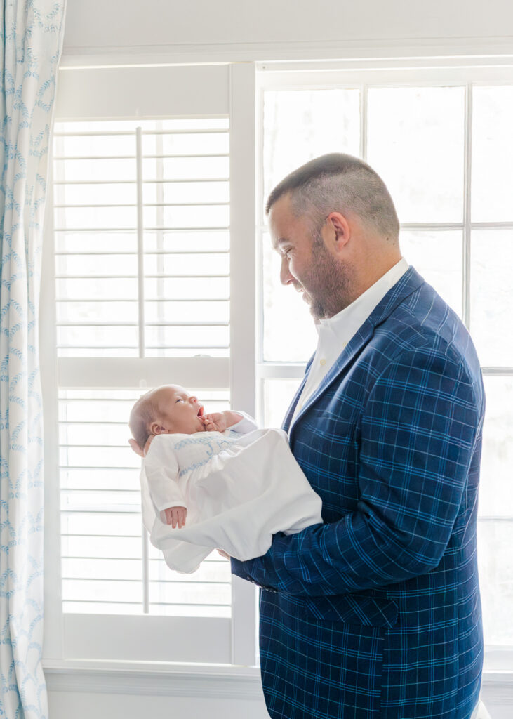 Dad standing by window holding newborn baby boy in white gown during newborn session at their home in Hoover, Alabama