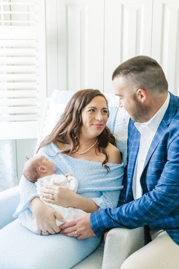 Mom holding sleeping newborn baby boy in rocker looking up at dad smiling lovingly at each other during in home newborn session with 5U Photography at their home in Hoover Alabama