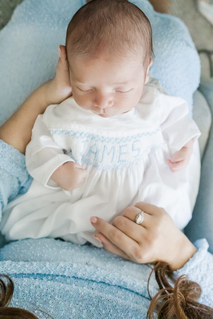 close up of baby boy is white gown with james smocked on the front in blue thread sleeping peacefully in mom's arms