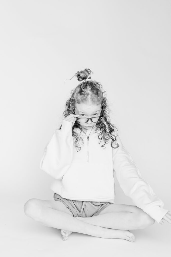 curly haired girl sitting with legs crossed on the ground looking over the top of glasses at camera during session with 5U Photography