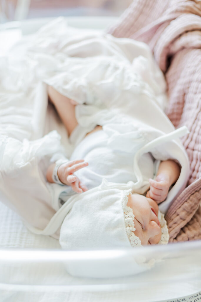 newborn baby girl laying in hospital crib wearing white shirt and bonnet during fresh-48 session 