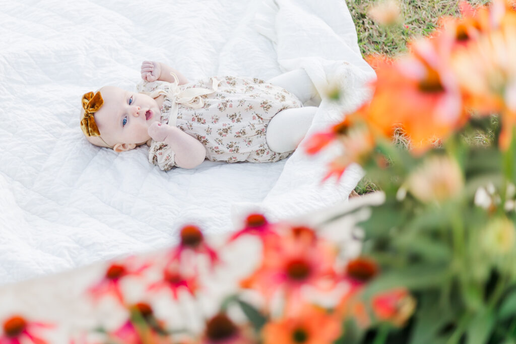 6 month old Charlotte laying on white blanket looking at beautiful flowers during 6 month milestone membership session at Old Baker Farm in Harpersville AL with 5U Photography