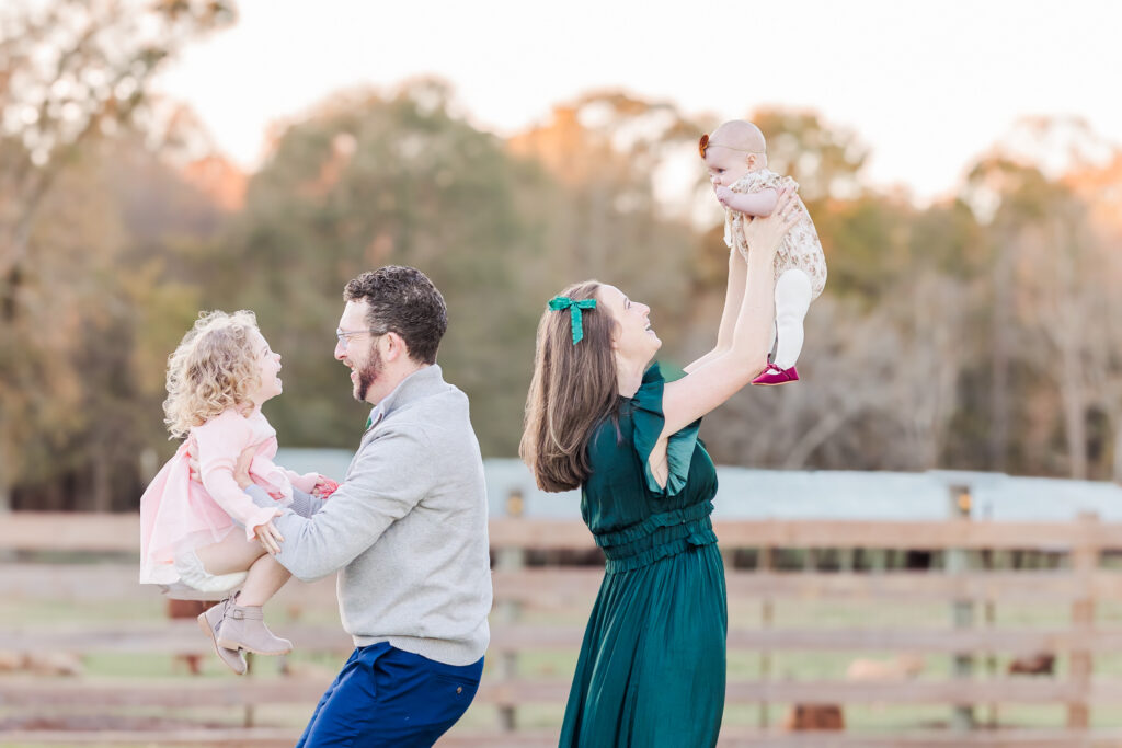 Mom and dad standing back to back holding up kids in the air as they giggle with joy during 6 month milestone session at Old Baker Farm in Harpersville Alabama