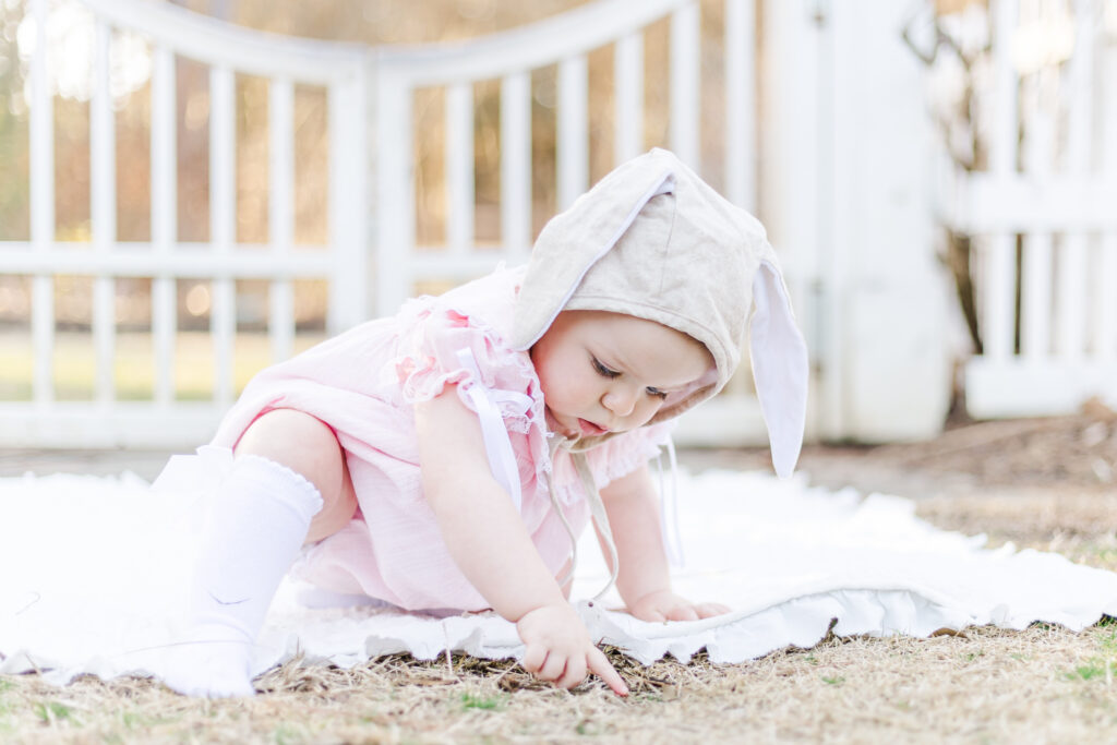 little girl with bunny bonnet on sitting on white blanket at the birmingham botanical gardens during 12 month milestone and smash cake session with 5U Photography