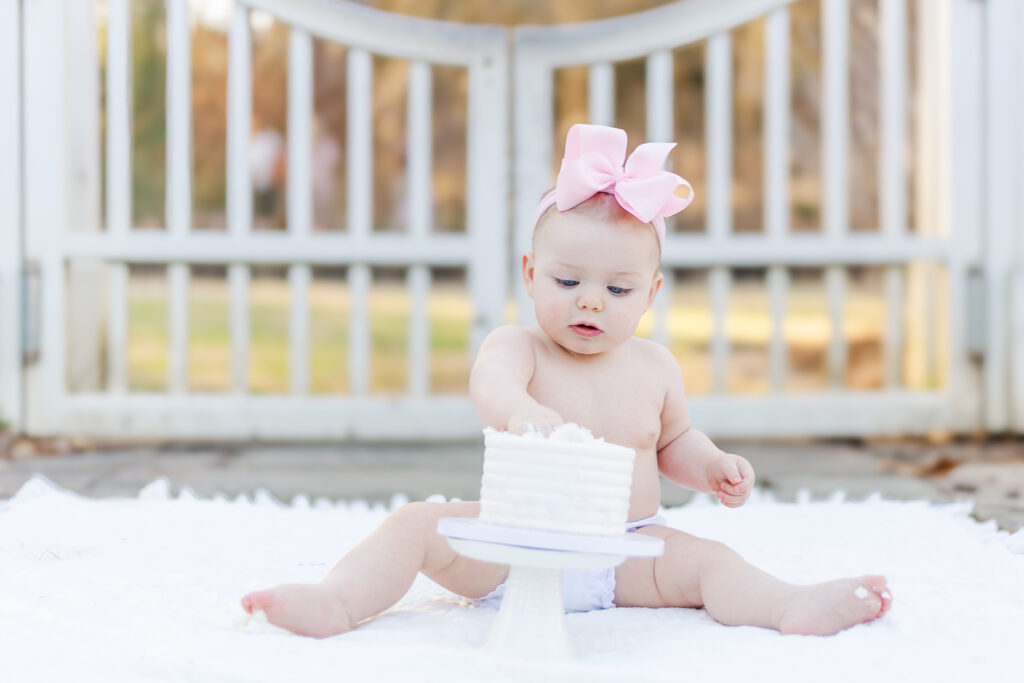 Little girl in pink bow and white bloomers sitting on white blanket during 12 month milestone and smash cake session at the birmingham botanical gardens