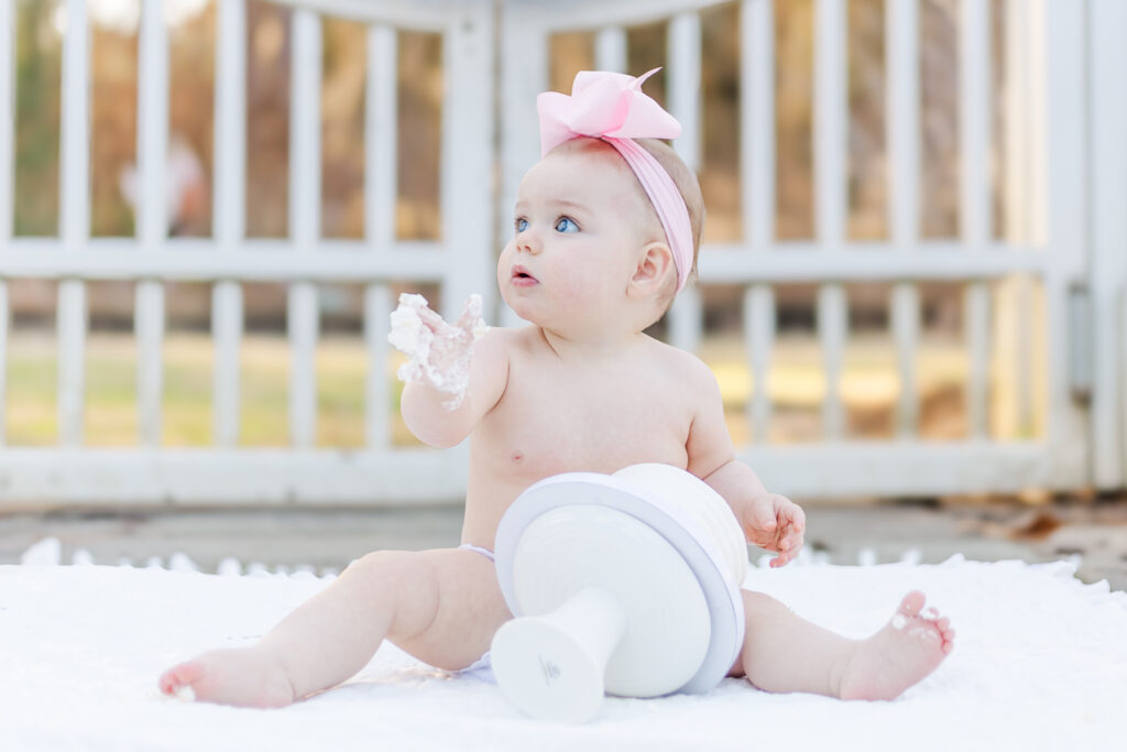 Baby girl playing with her smash cake in the beautiful gardens, with vibrant flowers in the background, captured by 5U Photography.