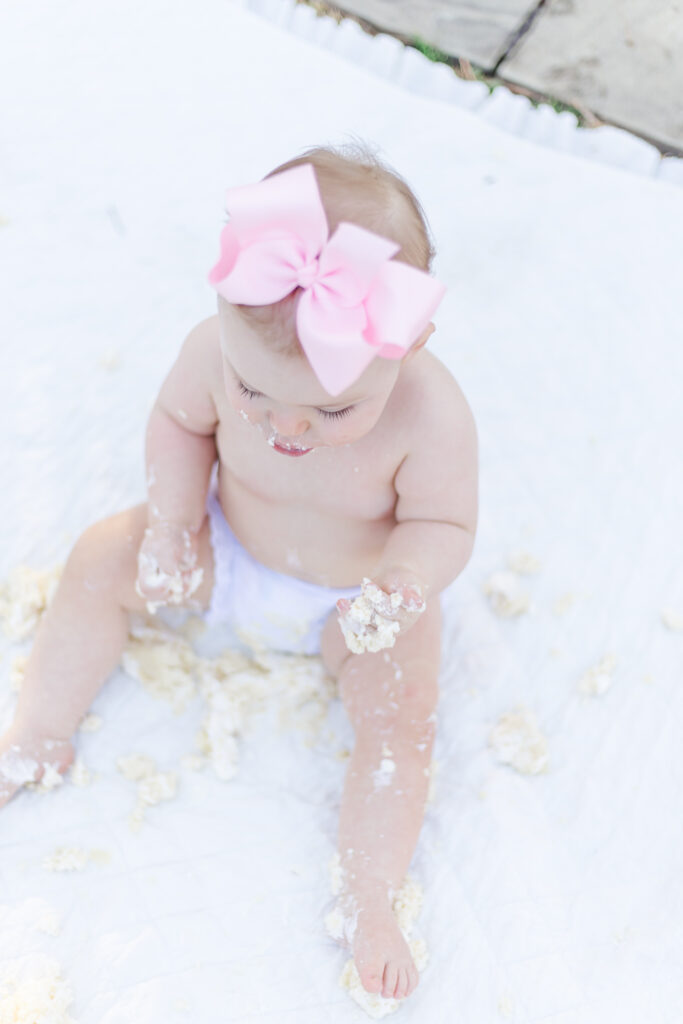 baby girl with frosting on hands and feet sitting on white blanket at the botanical gardens during 12 month milestone session