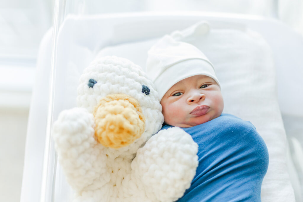 newborn baby boy laying in hospital crib  next to crochet duck lovey at Brookwood Hospital in Birmingham AL during fresh- 48 session with 5u photography