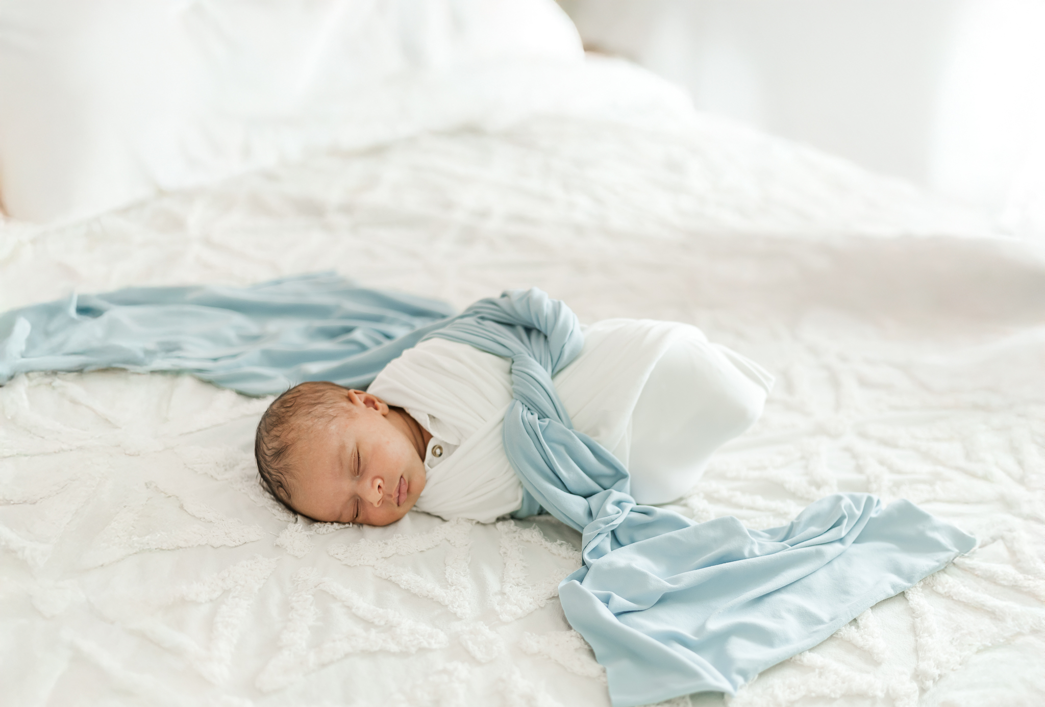 Newborn baby boy swaddled in a white blanket with baby blue sash sleeping peacefully on bed during in-home newborn session in birmingham, alabama