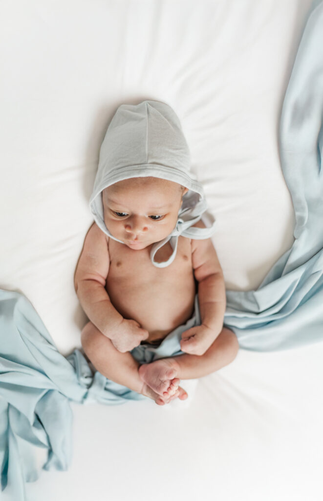 newborn baby boy lying on white blanket wearing a blue bonnet during in-home newborn session