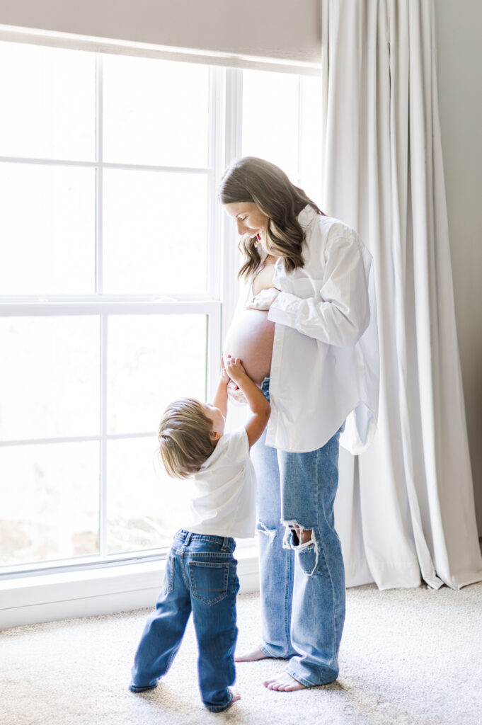 mom posing in front of large window in home in Birmingham AL with toddler son reaching up to touch her pregnant belly