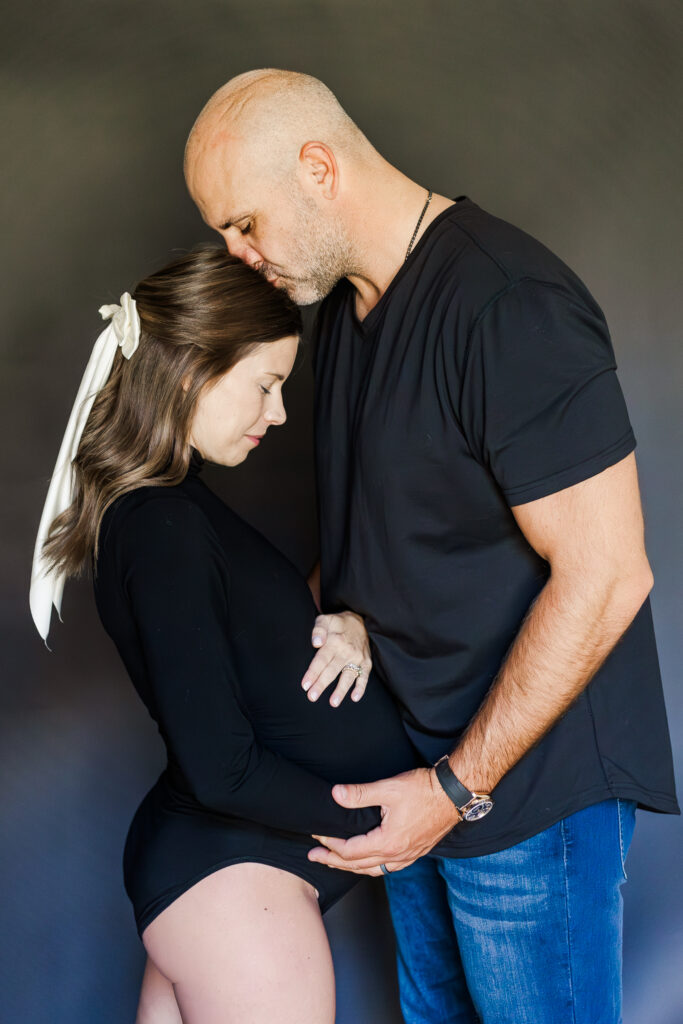 dad kissing mom on the head during in-home studio style maternity session in front of black seamless backdrop 