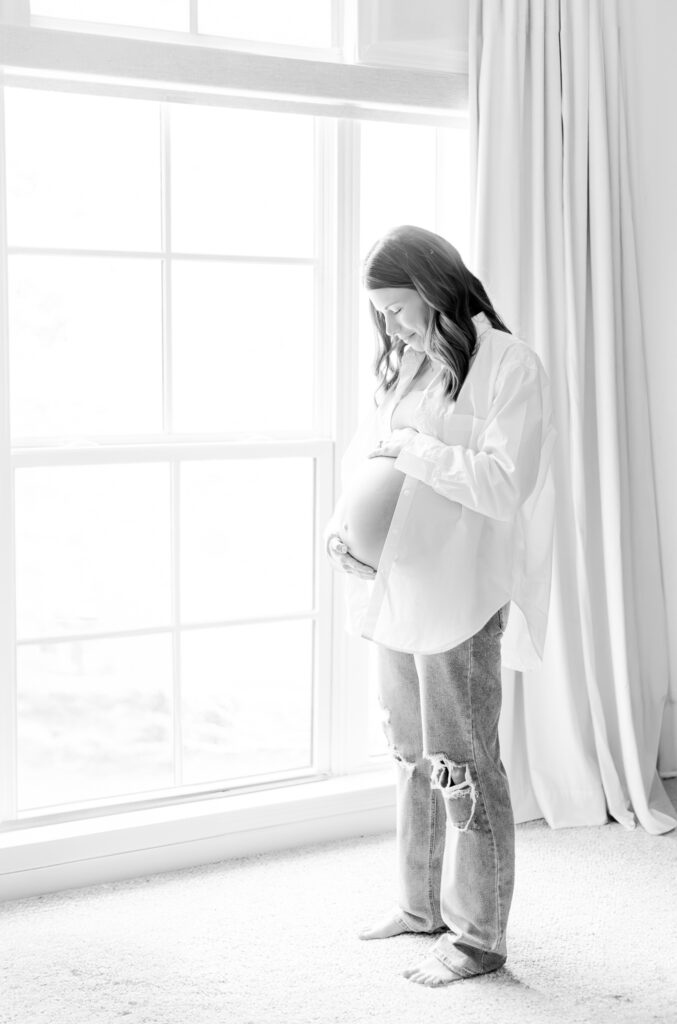 A cozy and intimate shot of the expecting mother standing in front of window in the nursery at home during in-home photo session with 5U Photography