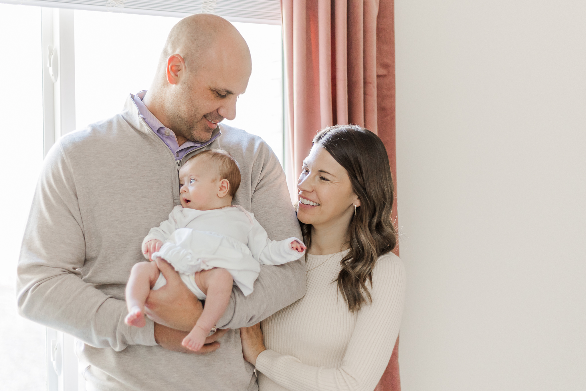 Mom and dad standing in front of window holding newborn baby girl during milestone membership in-home newborn session at their home in Birmingham, AL