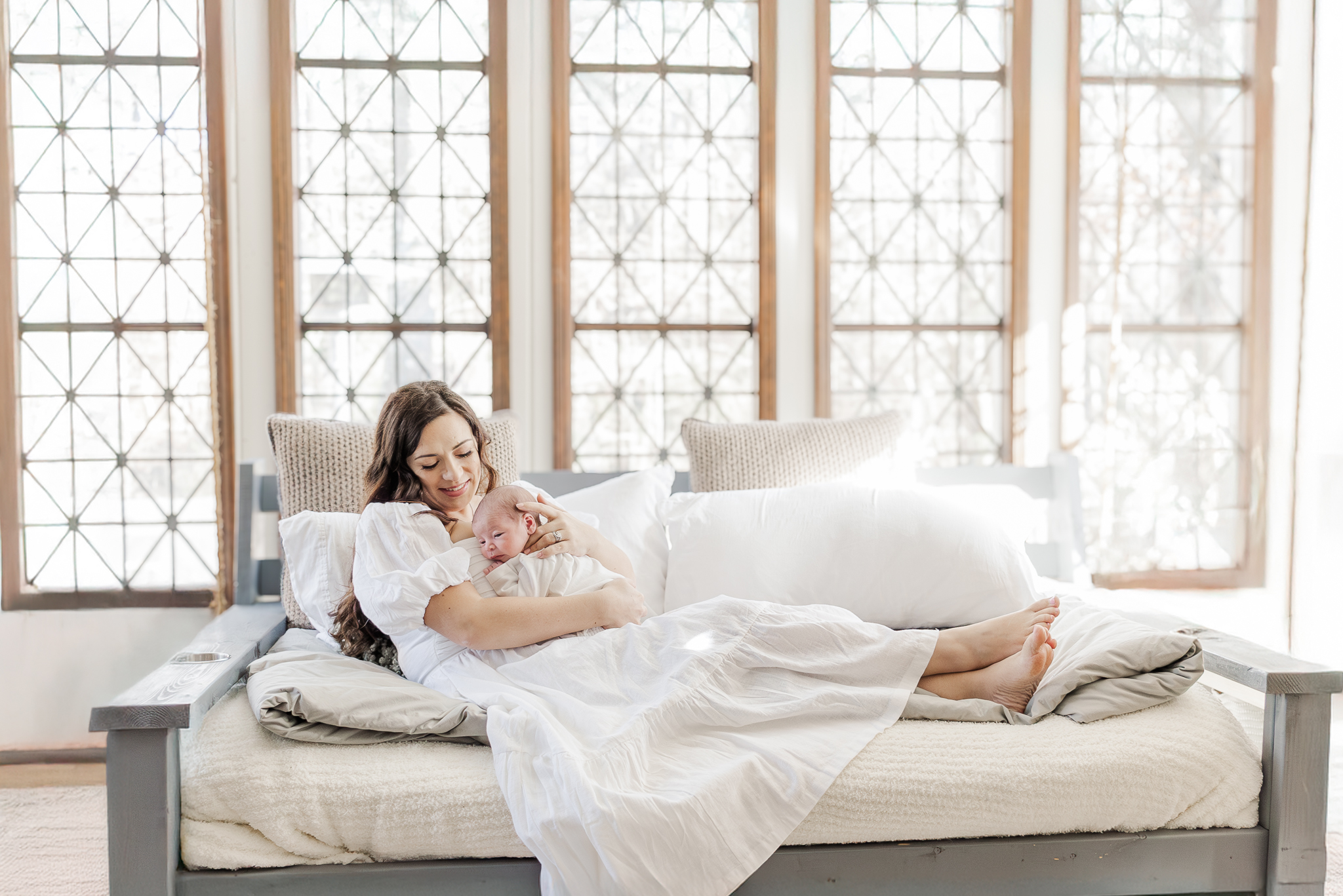 Mom laying on sunroom swing holding newborn baby boy swaddled in white blanket