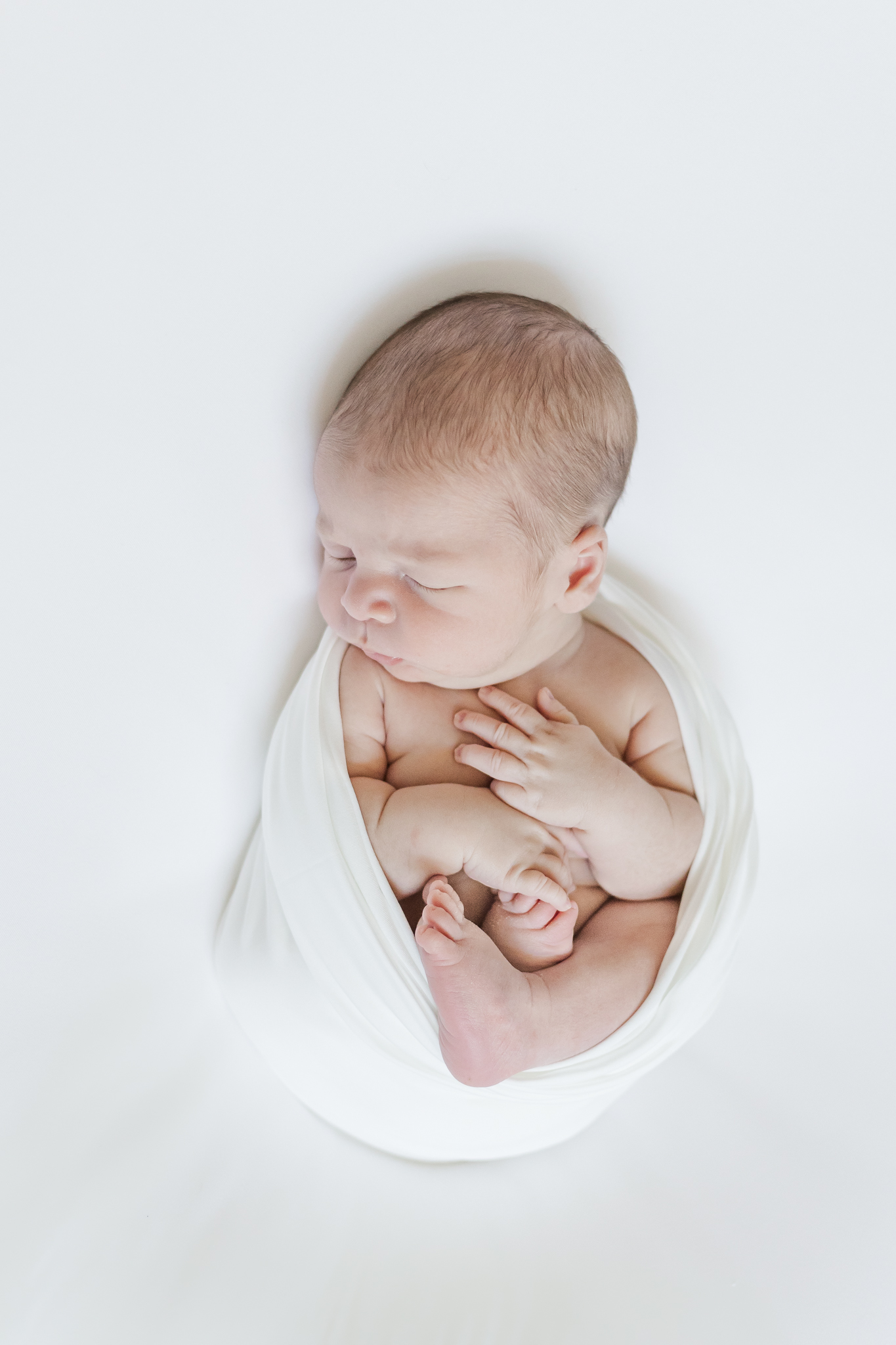 newborn baby boy laying on white blanket swaddled in white swaddle during in-home, lifestyle newborn session as a part of milestone membership