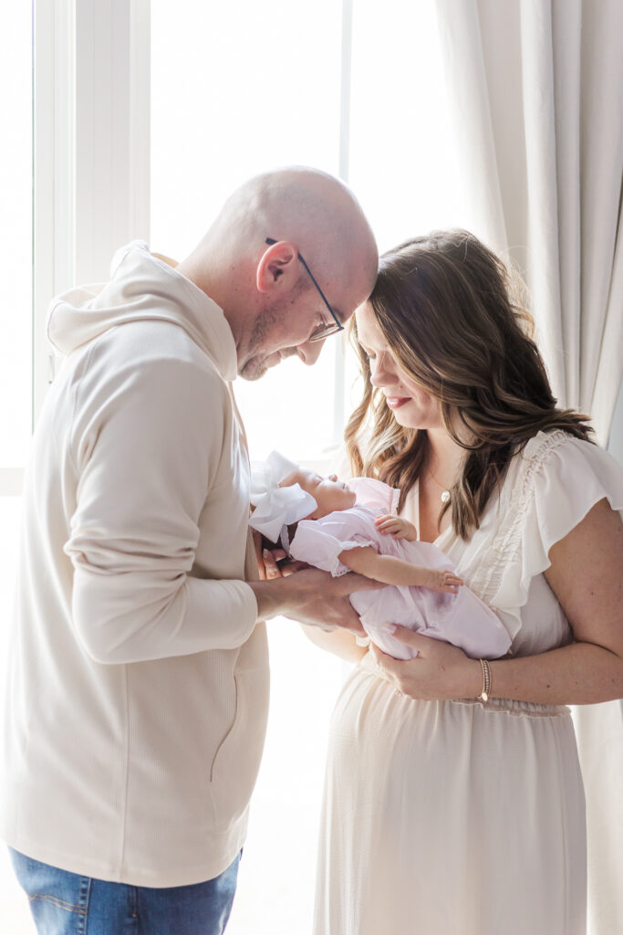 mom and dad holding new baby girl between them standing in front of window in their home in Gardendale, AL