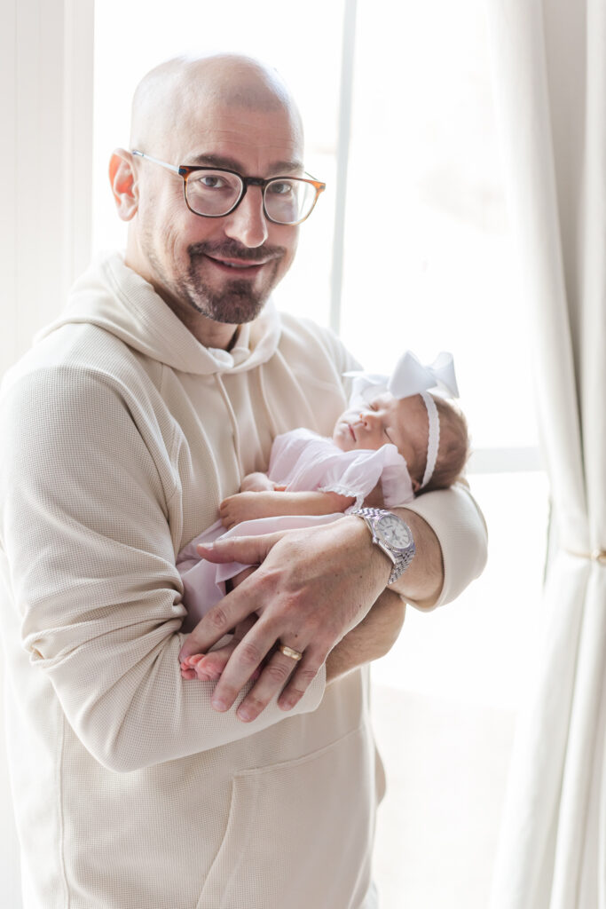 Dad standing in front of window holding newborn baby girl during session wtih 5U Photography