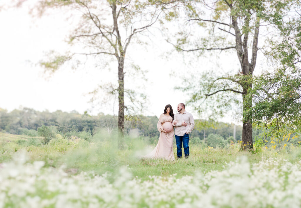 Expecting mother standing in field of flowers with her partner looking down and cradling her pregnant belly in sweet maternity session with 5U Photography