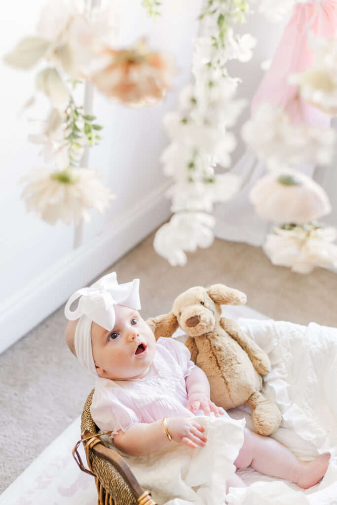 6 month old baby girl sitting in wicker basket surrounded by soft blankets looking up at beautiful flower mobile with cream and pink flowers during milestone and family session with 5U Photography