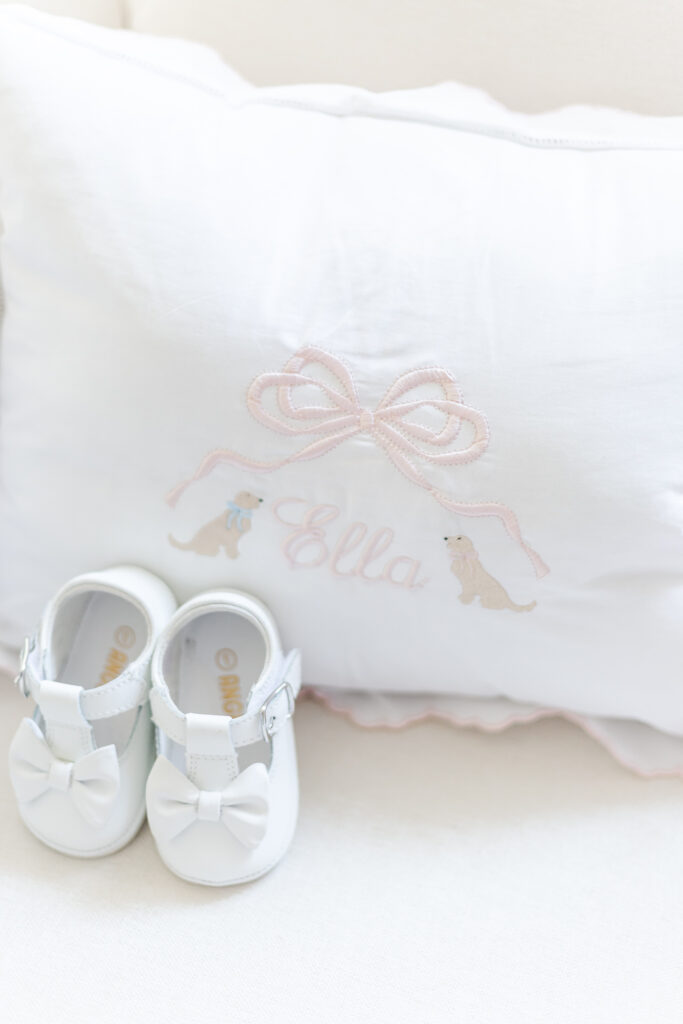 detail shot of embroidered pillow with name Ella in pink flanked by two golden retrievers with 1st pair of shoes propped in front
