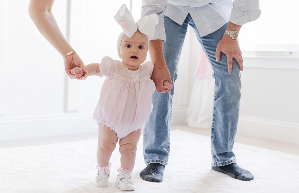 Ella standing up in her 1st pair of shoes holding mom and dad's hands showing off her newly achieved milestones during family session at their home in Calera AL