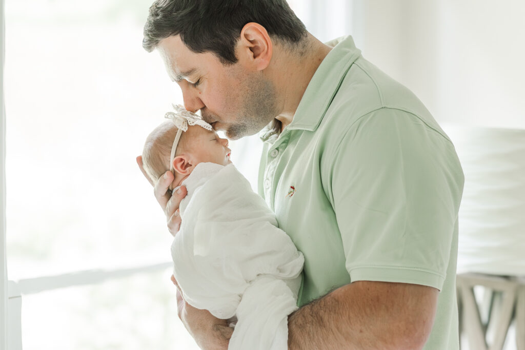 Father cradling newborn Eleanor in his arms while standing by a large window, gentle light illuminating the moment during in-home newborn session