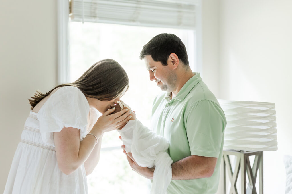 Dad holding Eleanor in front of window in parents room in Highland Lakes with mom kissing baby girl on the head while dad smiles down lovingly