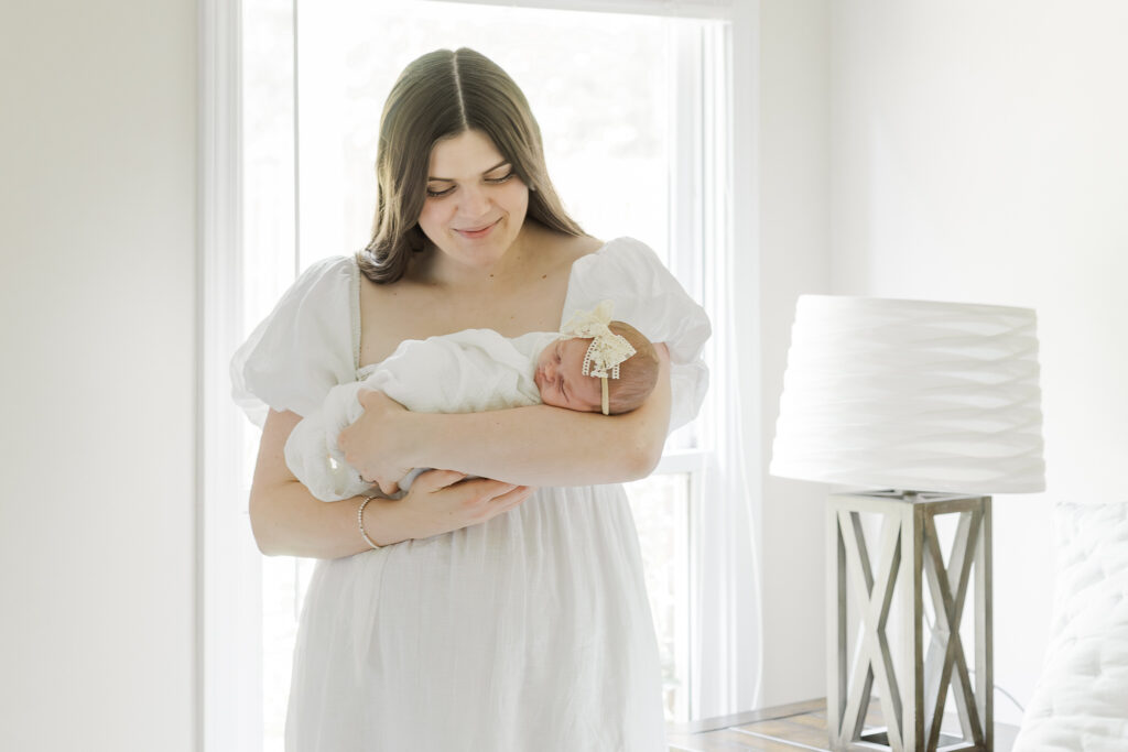 Mom standing in bedroom with natural light streaming in during in-home session cradling swaddled newborn baby girl