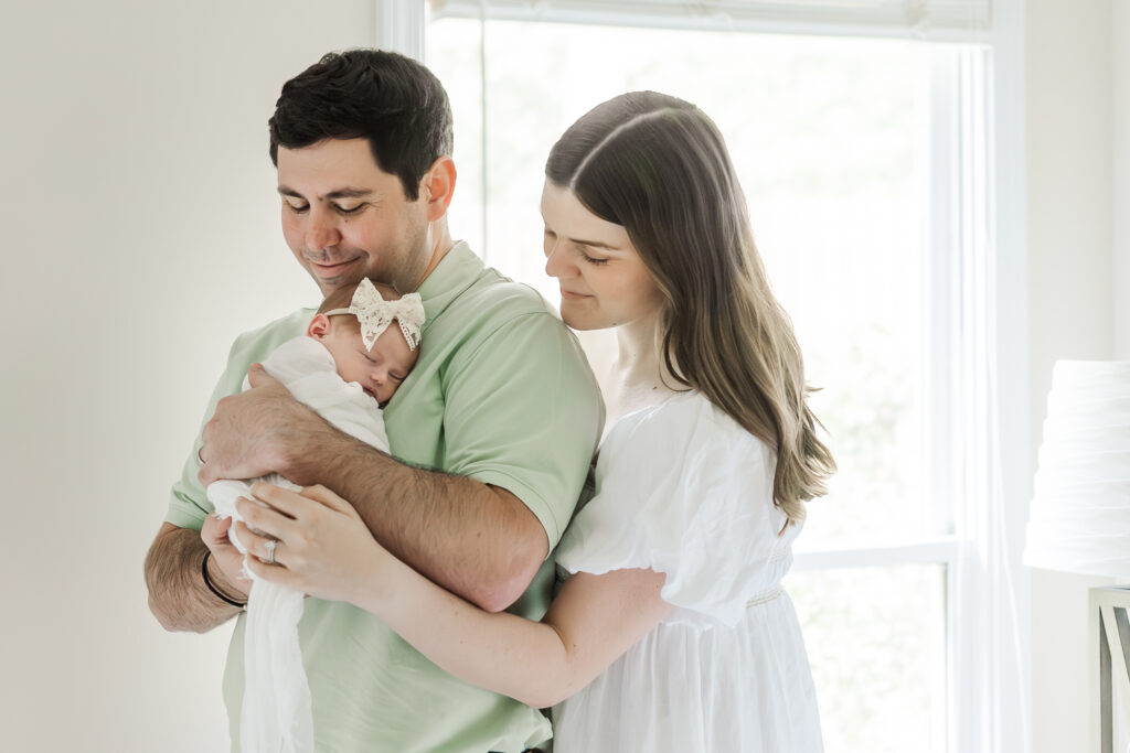 Mom and Dad standing next to their bed, with dad holding baby girl both smiling down at her with love and joy during in-home newborn session