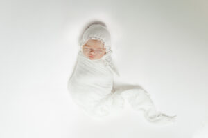 Eleanor swaddled in a white swaddle with matching white bonnet sleeping on a white backdrop during in-home session at family's home in highland lakes