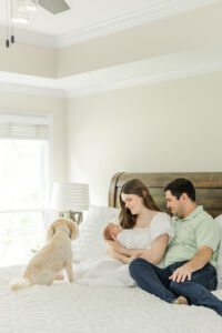 mom and dad sitting on bed holding newborn baby girl with white dog sitting  at their feet looking at new baby