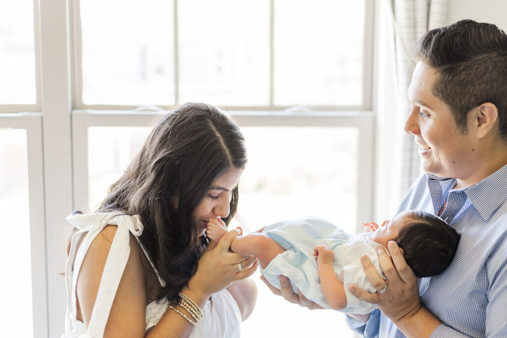 Parents smiling while mother kisses newborn’s feet near a sunlit window during an in-home newborn photography session in Trussville, Alabama.