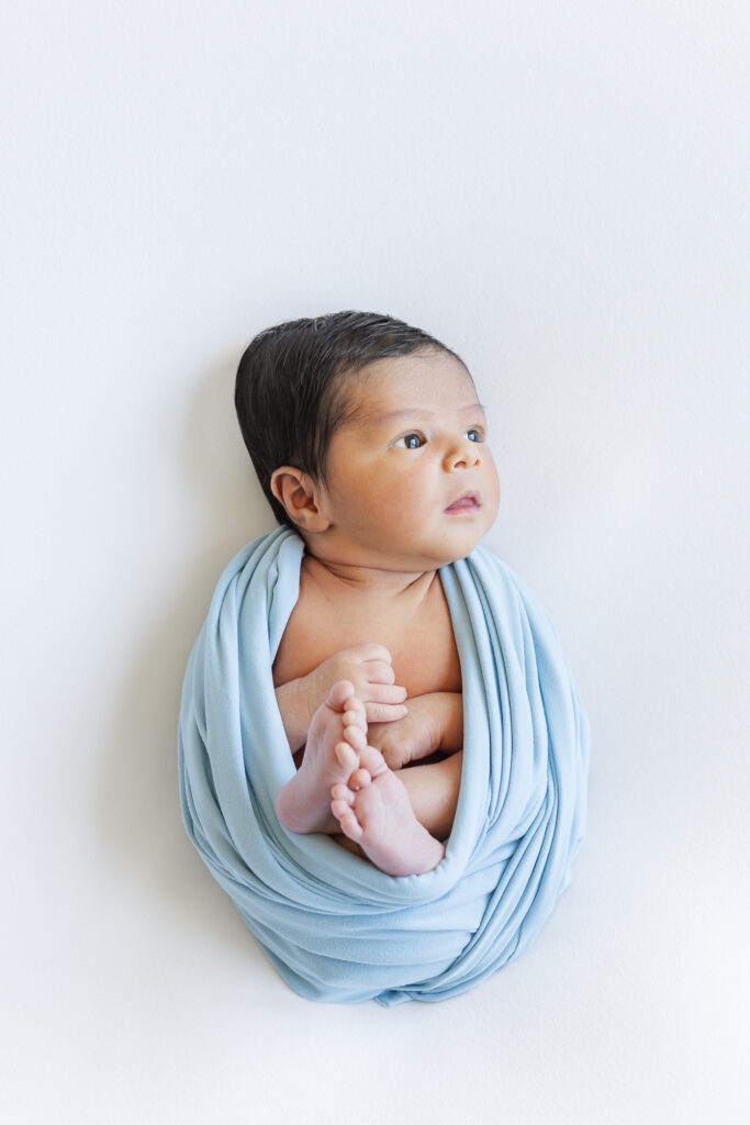Newborn baby boy wrapped in a soft blue swaddle, lying on white blanket during a Trussville, Alabama newborn photography session.
