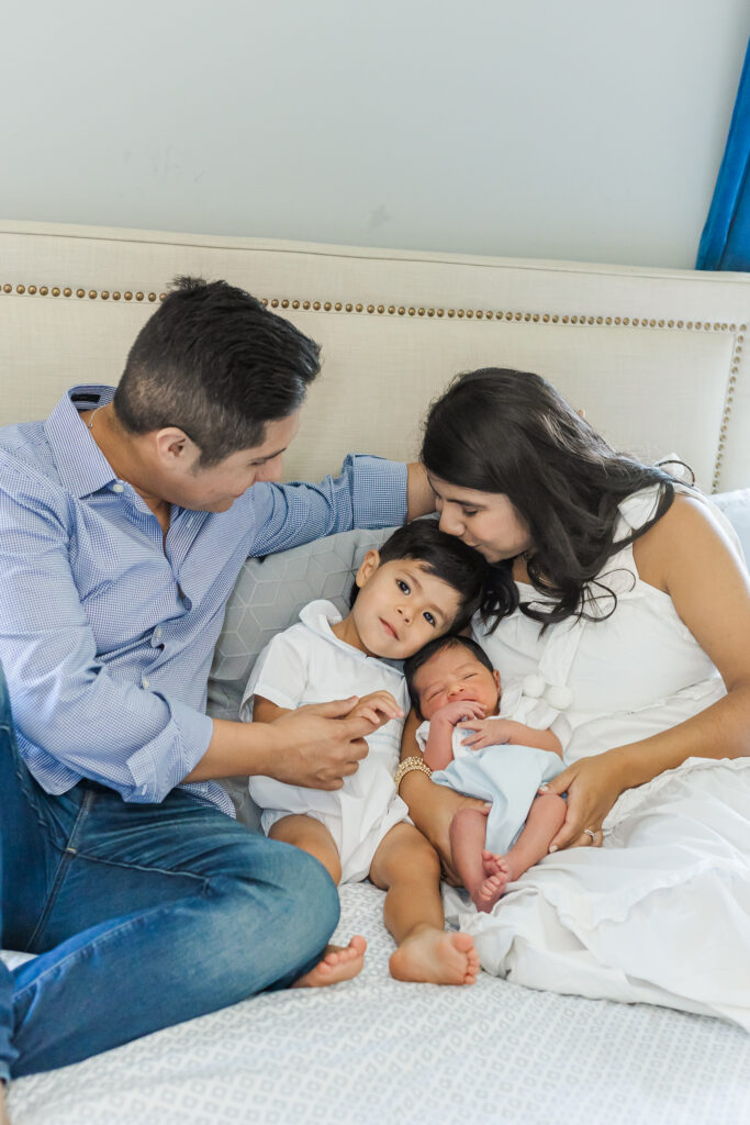 Mother kissing toddler while father holds newborn baby, all snuggled together on bed during a lifestyle newborn session in Trussville, Alabama.