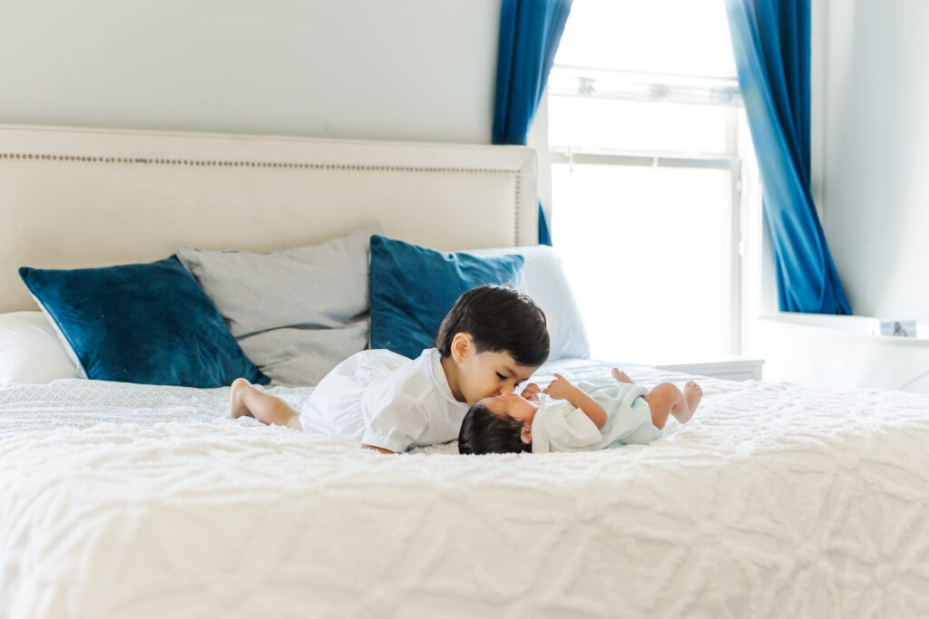 Toddler big brother giving newborn baby a forehead kiss while lying on bed during a Birmingham lifestyle newborn photography session.