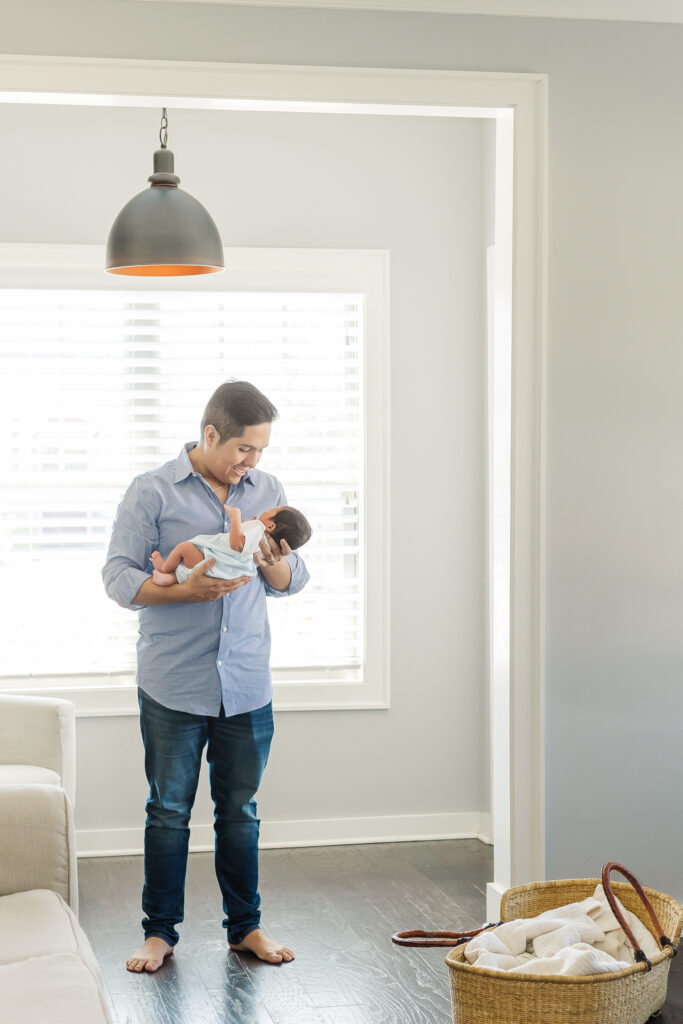 Father standing barefoot in living room holding newborn baby boy during a Trussville, Alabama lifestyle newborn photography session.