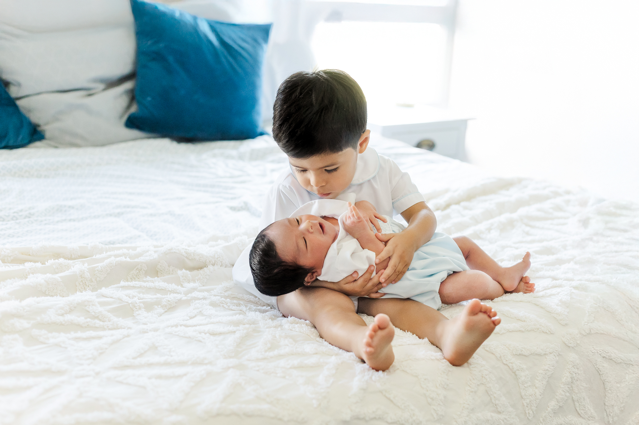 Sweet big brother holding and kissing newborn baby brother on the bed during a natural light in-home newborn photography session in Trussville, Alabama.