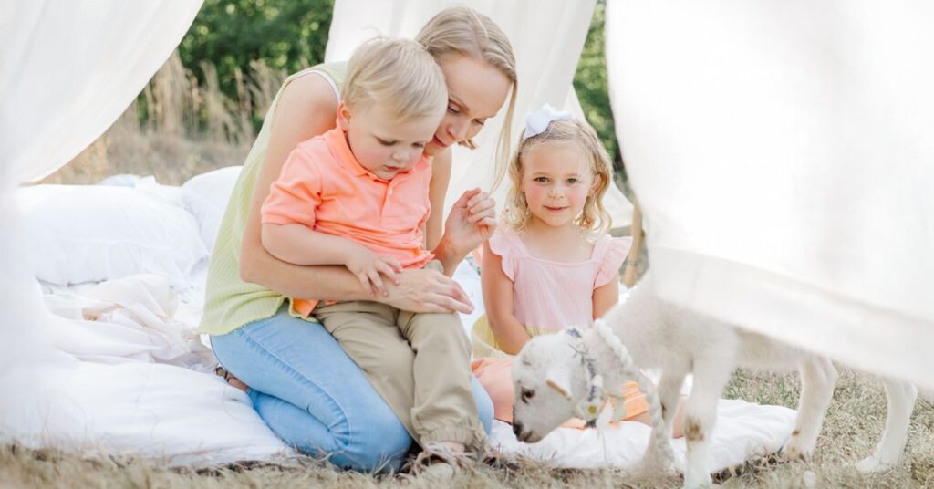 A mother sitting on white linens with her two young children as they gently watch a small lamb during a soft, heirloom-style Easter mini photo session outdoors.
