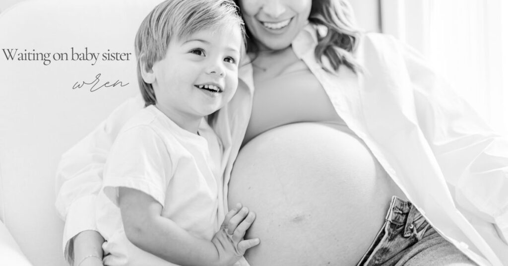 Black and white maternity portrait featuring a young child smiling while resting his hand on his mother’s baby bump as they wait for baby sister.