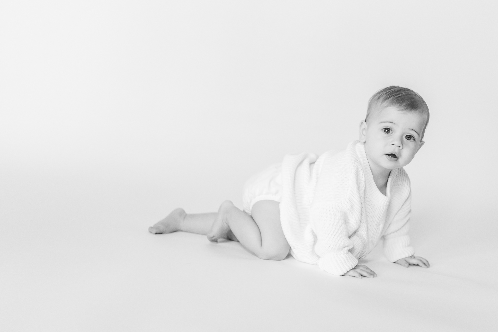 Black and white portrait of one year old baby girl crawling on a clean white backdrop during an in-home milestone session.