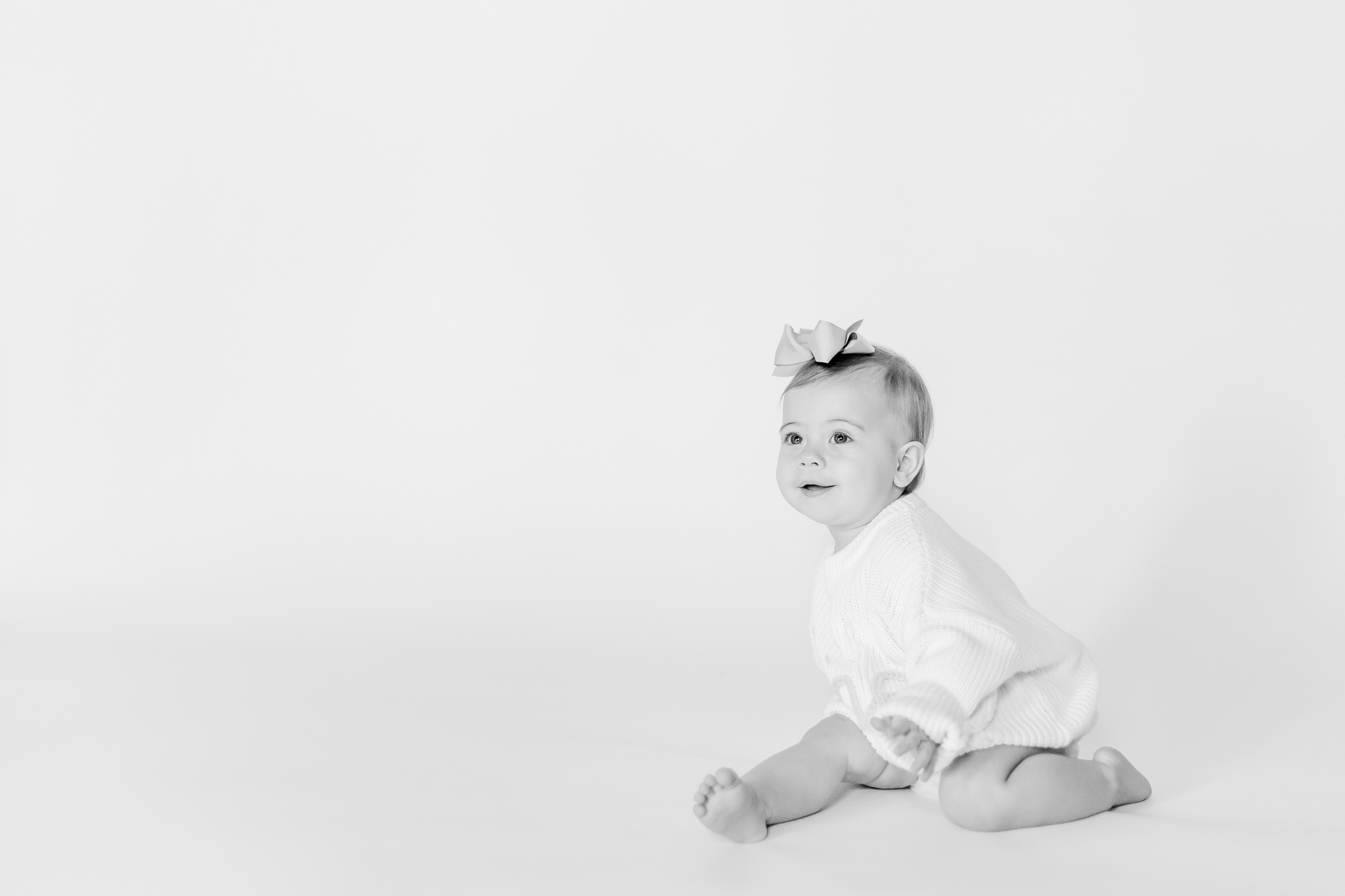 Heartfelt black and white portrait of one year old baby girl sitting on a white backdrop, wearing a soft sweater romper and bow